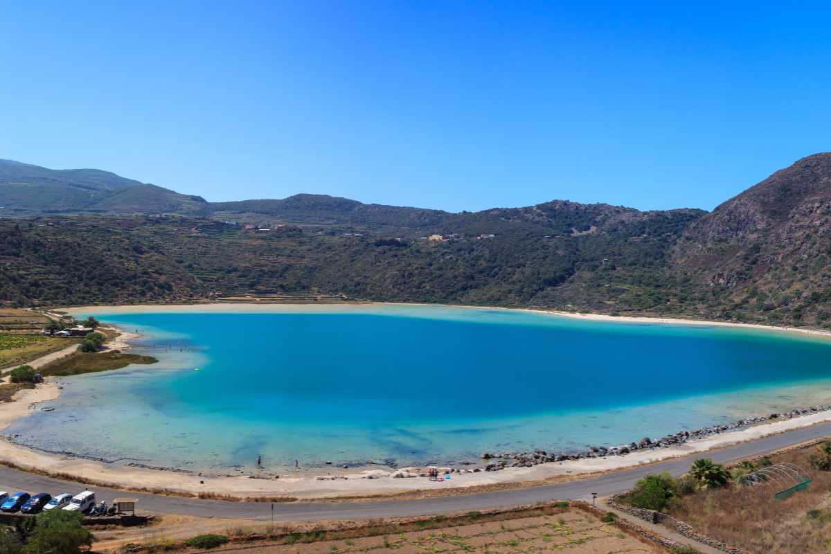 Veduta del lago Specchio di Venere, persone che si immergono nei fanghi, colline verdi e cielo riflesso sull'acqua, atmosfera rilassata tipica di Pantelleria